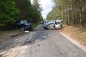 Na drodze asfaltowej policjant sporządza dokumentację. Droga z obydwu stron otoczona jest wysokimi drzewami. Po lewej stronie na poboczu widoczny prawy bok samochodu koloru granatowego z uszkodzonym przodem i  przednią szybą. Po prawej stronie, częściowo na jezdni, a częściowo na poboczu stoi pojazd koloru srebrnego. Bok i przód pojazdu jest uszkodzony, samochód ma rozbitą szybę. Drzwi boczne i tylne samochodu są otwarte.