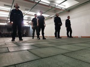 czterech policjantów stoi przodem na strzelnicy w rzędzie przed czerwoną linią. Mają opuszczone dłonie. Za nimi stoi instruktor.