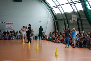 policjantka stoi obok dziewczyny, która na oczach ma alkogogle. Na hali sportowej rozstawione są żółte pachołki. Po prawej stronie siedzą dzieci