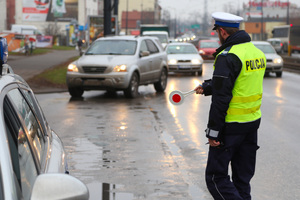 policjant ruchu drogowego na drodze daje znak tarczą do zatrzymania. Za nim widoczny pojazd