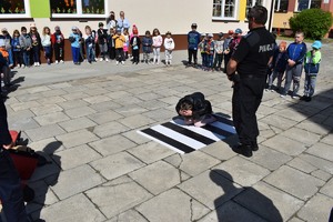 Dziewczynka demonstruje na macie bezpieczną pozycję żółwia. Policjant stoi obok niej. Obserwatorami są dzieci