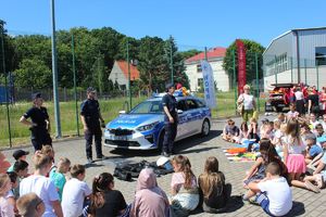 policjanci przy radiowozie prowadzą pogadankę, w koło siedzą uczniowie