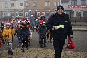 dzieci w mikołajkowych czapkach idą z policjantem trzymającym odblaski i worek świętego Mikołaja