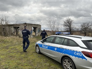 Policjanci idą w kierunku pustostanu. Na pierwszym planie widać radiowóz policyjny.