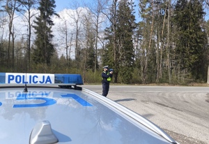 Policjant stoi na drodze i mierzy prędkość. Na pierwszym planie widać radiowóz.